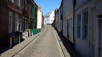 This urban photograph shows a quiet Whitby street in Yorkshire during the afternoon in spring. The cobbled road is lined with traditional brick and painted terrace houses that reflect the architectural character typical of Whitby. Bright sunlight highlights the facades and creates contrasting shadows along the pavement. The street gently curves upward, leading the eye toward more houses in the distance under a clear blue sky, capturing the peaceful atmosphere of this historic Yorkshire coastal town.